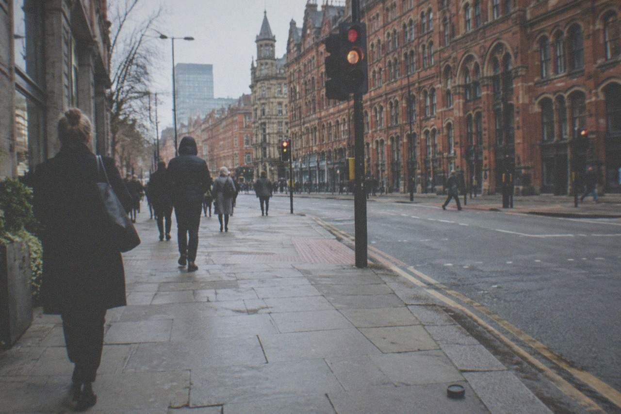 Pedestrians walking along a busy Manchester city centre street with red brick Victorian buildings above them