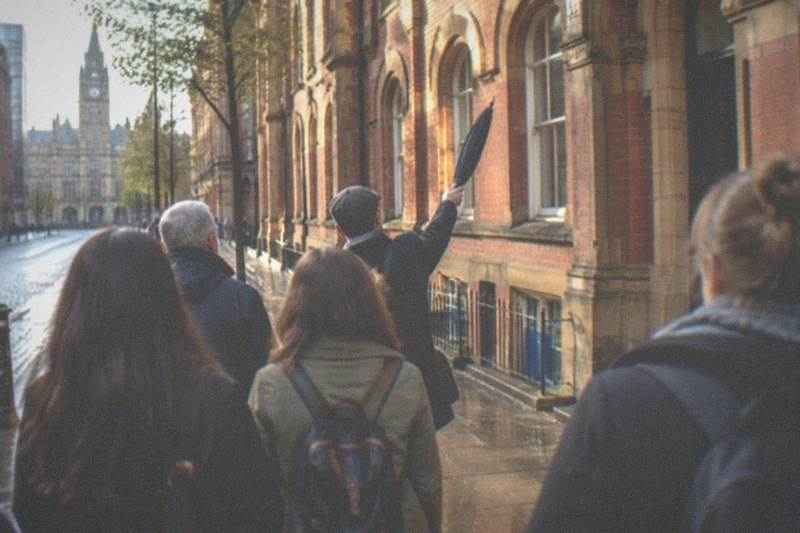 A guide leading a small free walking-tour group past a red-brick Victorian terrace in Manchester city centre on a bright afternoon.